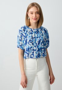 Giovane donna con capelli biondi fino alle spalle, che indossa una blusa blu a motivi e pantaloni bianchi, in piedi davanti a uno sfondo chiaro e uniforme.