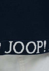 Μάλλινο ρεβέρ σε ναυτικό μπλε με λευκούς έντονους χαρακτήρες "JOOP!" πάνω από ανοιχτό μπεζ ύφασμα.