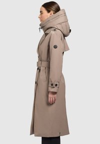 Trench-coat long beige avec une grande capuche matelassée, taille ceinturée, poignets à boutons et un écusson logo sur la manche. Texture du tissu lisse.