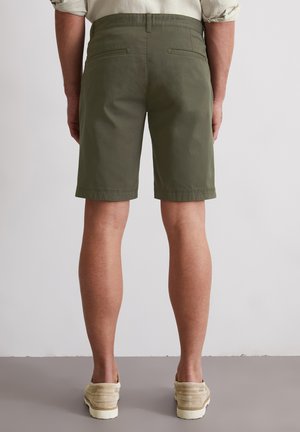 Olivengrüne Shorts aus Baumwollstretchmaterial mit aufgesetzten Taschen auf der Rückseite. Der Saum sitzt knapp über dem Knie und wird mit beigen Schuhen kombiniert.