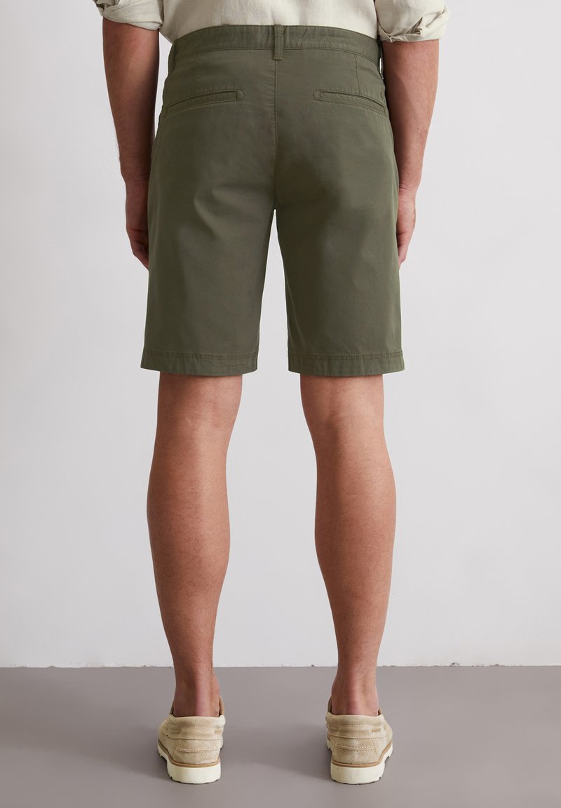 Shorts vert olive en tissu coton extensible, avec poches arrière. L'ourlet se situe juste au-dessus du genou, associés à des chaussures beiges.