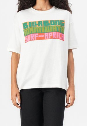 Henkilö, joka käyttää valkoista T-paitaa, jossa on värikästä retrotyylistä tekstiä "Billabong Mami Wata Surf-Africa" rintamuksessa, yhdistettynä mustiin housuihin.