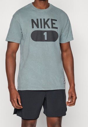 Mann trägt ein blaues Nike T-Shirt mit „NIKE 1“-Logo und schwarze Shorts, steht vor einem schlichten weißen Hintergrund.