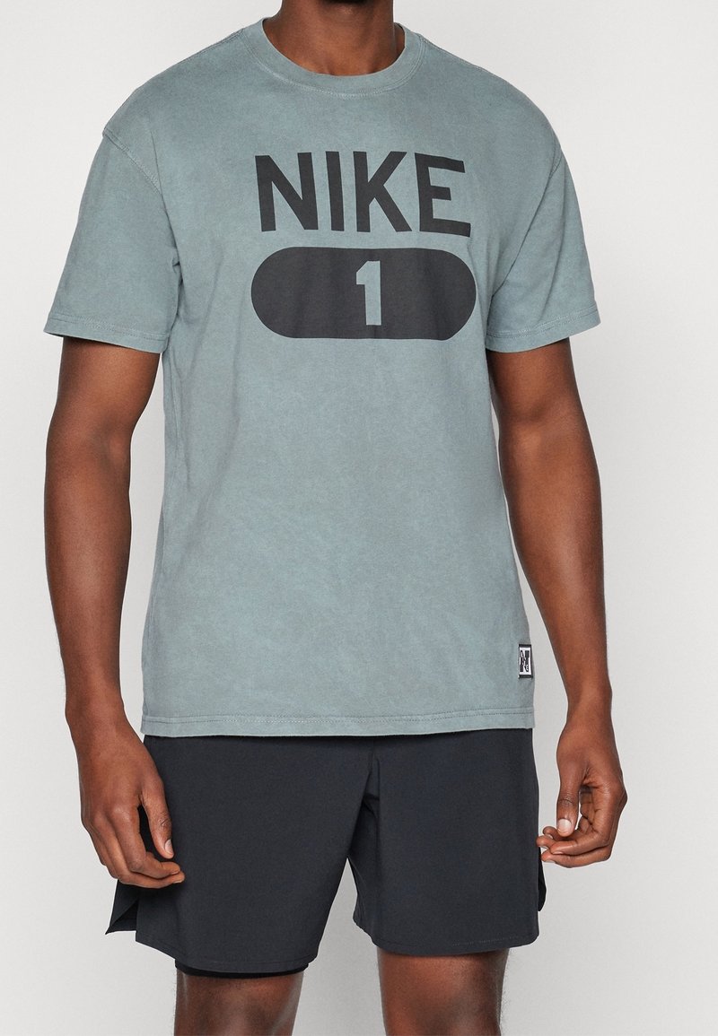 Homme portant un t-shirt bleu Nike avec le logo "NIKE 1" et un short noir, debout devant un fond blanc uni.