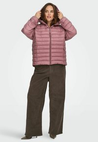 Kvinne som justerer hetten på en mosegrønn, vattert jakke, iført vide brune bukser og spisse brune sko, stående foran en ensfarget bakgrunn.