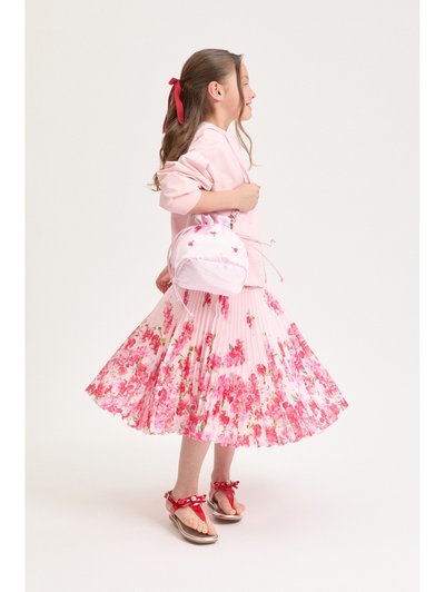 Νεαρή κοπέλα με ροζ πουλόβερ, φλοράλ πλισέ φούστα, κόκκινα σανδάλια και κορδέλα μαλλιών που ταιριάζει, κρατώντας μια μικρή ροζ τσάντα με κορδόνι.