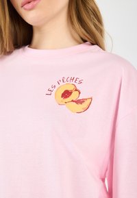 Světle růžové bavlněné tričko s grafikou dvou půlek broskví a textem "LES PÊCHES," s volným střihem a klasickým kulatým výstřihem.