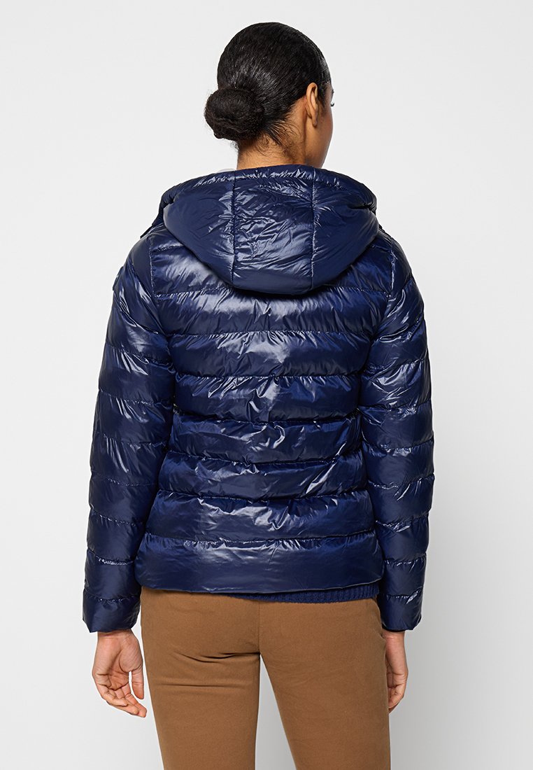 Eine Frau mit dunklem Haar im Dutt trägt eine glänzende marineblaue Kapuzenjacke und braune Hosen. Sie wird von hinten vor einem schlichten Hintergrund gezeigt.