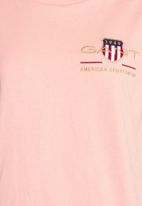 T-shirt rose clair avec logo brodé représentant un écusson, "GANT" en doré, et "AMERICAN SPORTSWEAR" en rouge en dessous.