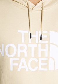 Béžová mikina s velkým bílým logem "The North Face", šňůrkami s černými konci a měkkou, texturovanou tkaninou.