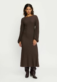 Kvinde iført en struktureret brun midi-kjole med lange ballonsleeves og mørkebrune knæhøje støvler med hæl, stående mod en ensfarvet baggrund.