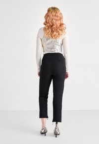 Metalický stříbrný cropped vest s žebrovanými dlouhými rukávy, v kombinaci s černými priléhavými kalhotami a stříbrnými podpatky, ukazující viditelnou protézu nohy.