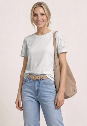 Uśmiechnięta kobieta z blond włosami związanymi do tyłu, ubrana w biały, wzorzysty top z krótkim rękawem, jasnoniebieskie dżinsy, beżowy pasek i beżową torbę na ramię.