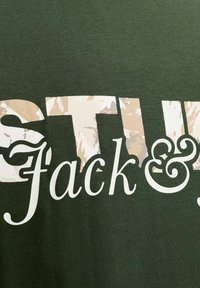 Camiseta verde con texto grande y llamativo. La palabra "STU" en blanco y "Jack &" en un patrón de camuflaje, con tela suave y texturizada.