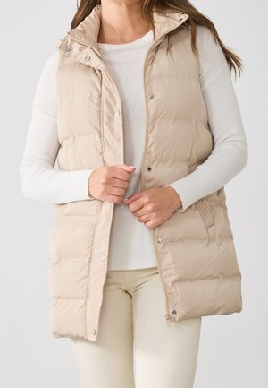 Mujer con un chaleco acolchado sin mangas beige sobre una camiseta blanca de manga larga con textura de canalé, abrochando la cremallera y los botones de presión del chaleco.