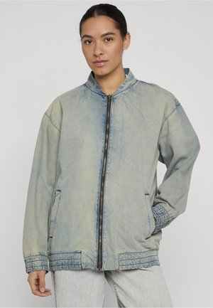 Jeune femme aux cheveux foncés portant une veste en denim bleu clair délavé avec fermeture éclair et un pantalon gris clair, debout avec une main dans la poche.