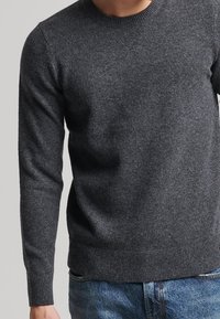 Mann in einem figurbetonten dunkelgrauen Langarmstrickpullover und blauen Jeans, der vor einem schlichten hellgrauen Hintergrund steht.