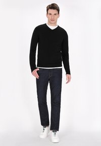 Maglione v-neck nero, maglietta bianca sotto, jeans blu scuro e sneaker bianche. Il maglione ha un design aderente e una texture liscia.