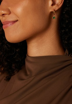 Créole en or avec une gemme verte suspendue en bas, présentée sur un cou lisse et brun et un haut marron.