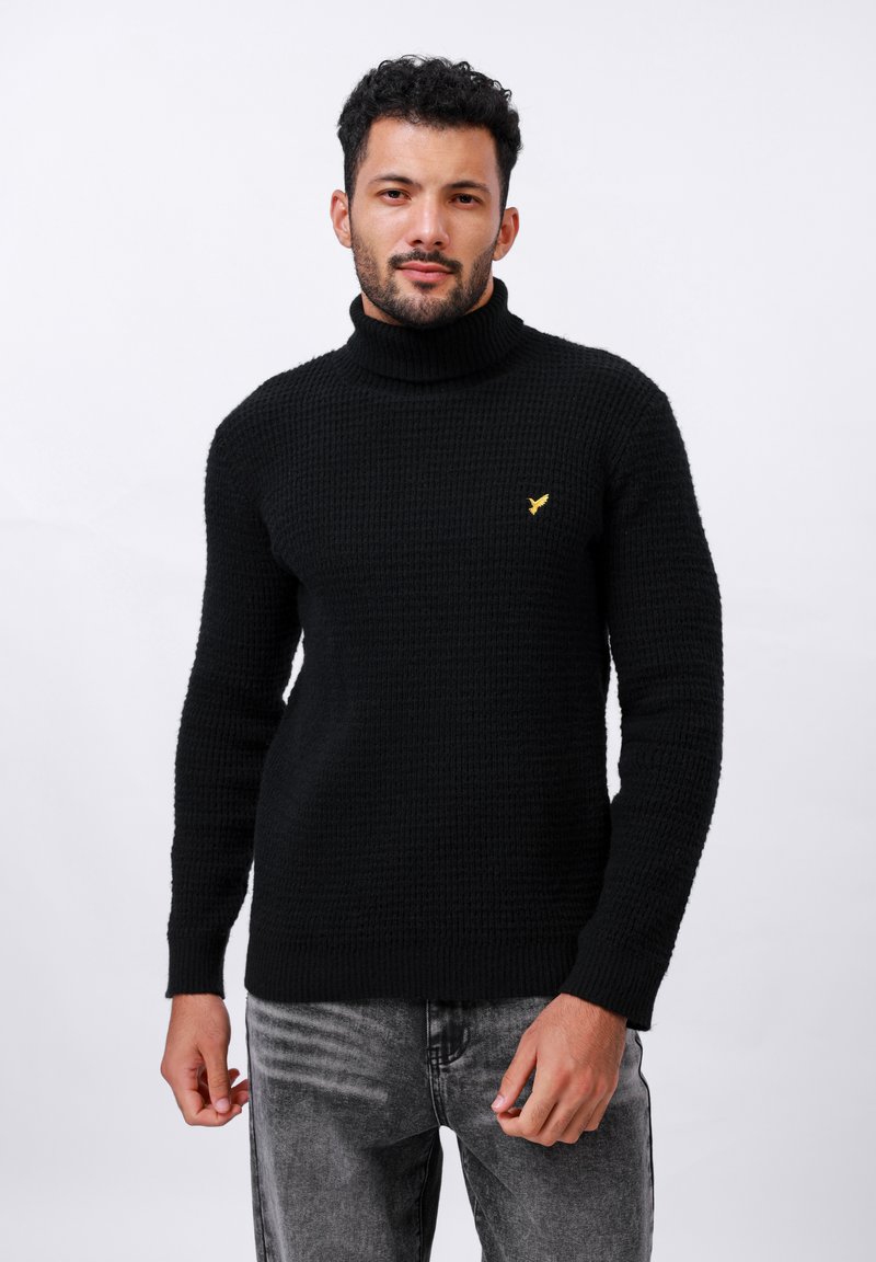 Maglione nero a collo alto in maglia con motivo testurizzato, dotato di un piccolo logo giallo sul lato sinistro del petto. Abbinato a jeans grigi.