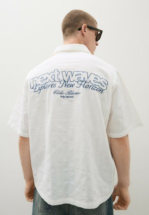 Mężczyzna w czarnych okularach przeciwsłonecznych i białej koszuli z krótkim rękawem z niebieskim napisem "next waves Explores New Horizon Côte Bleue" na plecach.