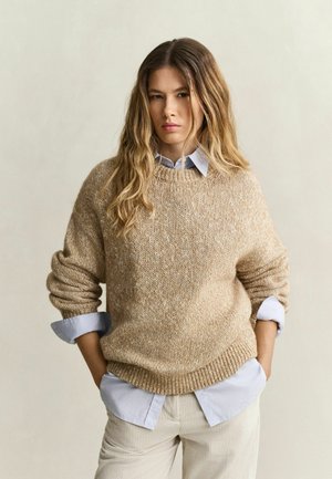Jeune femme aux longs cheveux ondulés portant un pull en maille beige sur une chemise bleu clair à col et un pantalon clair, les mains dans les poches.