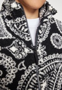 Giacca in pile bianca e nera con colletto alto, chiusura con zip e una trama intricata. Presenta colori a contrasto e una sensazione morbida.