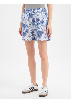 Vrouw draagt witte shorts met blauwe tropische bloemenprint en witte sneakers met zwarte strepen.