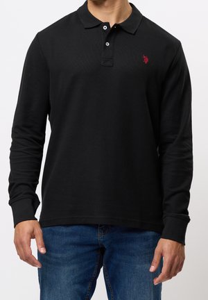 Mann trägt ein schwarzes Langarm-Poloshirt mit rotem Logo auf der Brust und blaue Jeans vor einfarbigem Hintergrund.
