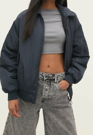 Femme portant une veste bomber bleu marine foncé par-dessus un t-shirt court gris et un jean gris taille haute acid-wash, avec une main dans la poche.