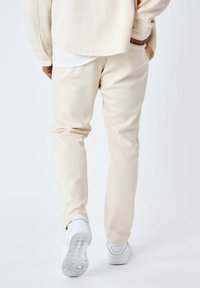 Beige joggers hebben een gladde textuur en een taps toelopende pasvorm, gecombineerd met witte sportschoenen met een rubberen zool en een gestructureerd profiel.
