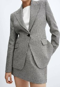 Femme portant un blazer pied-de-poule noir et blanc et une mini-jupe assortie avec un top blanc, les mains détendues sur les côtés et dans la poche.