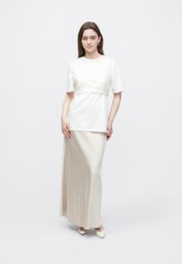Biele tričko s textúrovanou strednou časťou, skombinované s dlhou, prúdiacou béžovou saténovou sukňou. Modelka stojí pred jednoduchým pozadím.