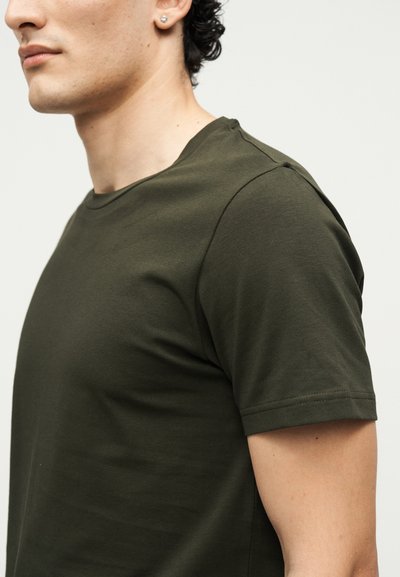 Νέο άτομο που φοράει ένα απλό σκουροπράσινο κοντομάνικο μπλουζάκι, εμφανιζόμενο από το πλάι σε φωτεινό φόντο.