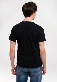 Mann mit dunklen Haaren, der ein einfaches schwarzes T-Shirt und eine blaue Jeans trägt, mit dem Rücken zur Kamera vor einem schlichten weißen Hintergrund.