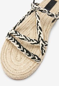 Gevlochten sandalen met een natuurlijke touwbasis. De banden zijn zwart en wit, wat een geweven patroon creëert. De textuur is glad met een plat ontwerp.