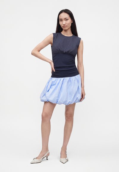 Femme debout, main sur la hanche, portant un haut sans manches bleu marine, une jupe boule bleu clair et des chaussures blanches à petit talon sur fond blanc.
