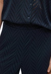 Tumši zila apģērba komplekta apakšējā daļa ar perforētu rakstu un ševronu dizainu. Augšdaļai ir pogu aizdare un brīvs fasona, savukārt tā ir apvienota ar augstām biksēm.