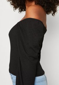 Mustat olkaimeton paita, jossa on teksturoitu kangas, pitkät hihat ja kapea siluetti, yhdistettynä vaaleansinisiin farkkuihin.