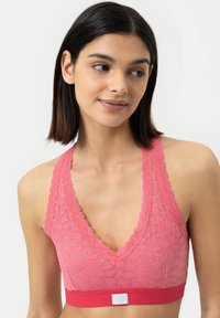Růžový krajkový bralet s hlubokým V výstřihem, zvlněnými okraji a širokým červeným pásem na spodní části s bílou logo štítkem.