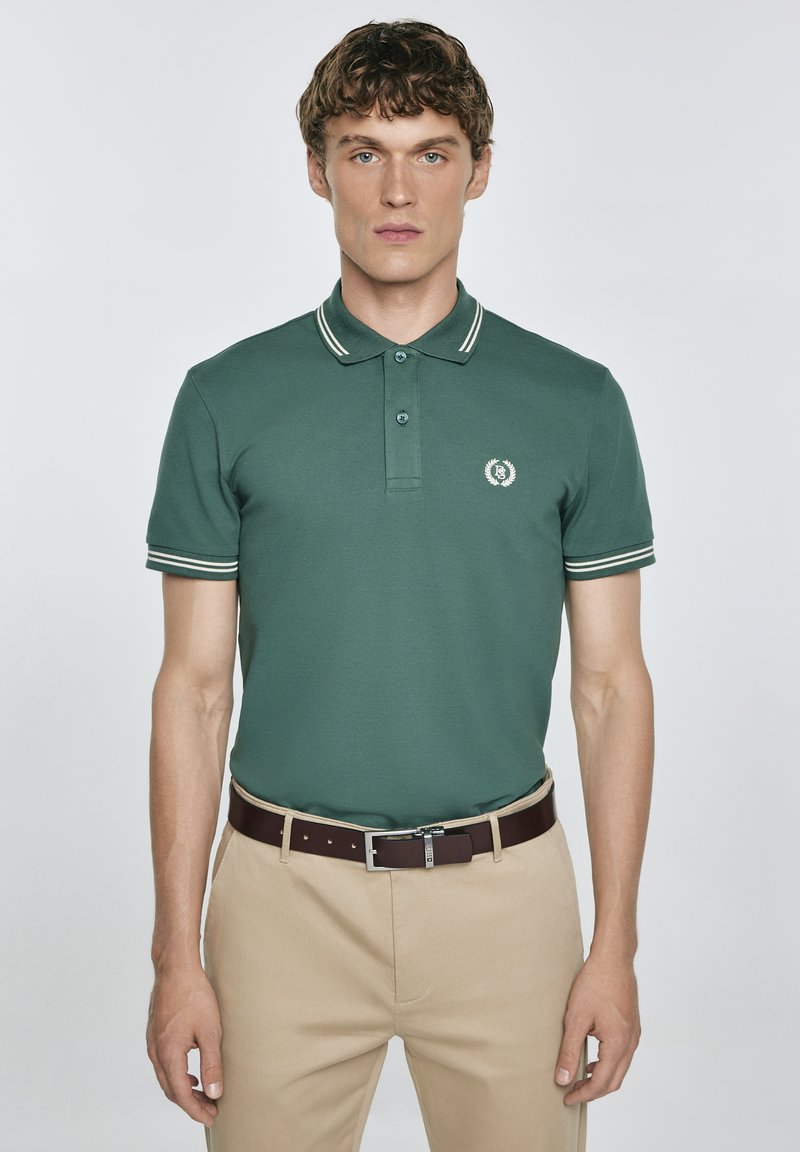 Zelená bavlněná polo košile s klasickým límcem, bílými pruhy a malým kulatým logem. V kombinaci s béžovými kalhotami a hnědým páskem.