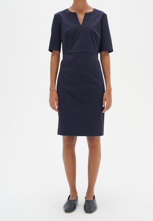 Woman wearing a knee-length, short-sleeve navy dress with a V-neckline and black slip-on shoes, standing against a plain light background.