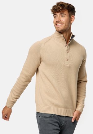 Suéter de punto beige con cuello alto, tres botones, puños y dobladillo con canalé, que presenta una textura suave y un diseño ajustado.