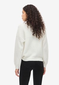 Bela pletena pulover s rebrasto teksturo na zgornjem hrbtu in balonastimi rokavi. Kratek dizajn, v kombinaciji s črnimi oprijetimi kavbojkami.