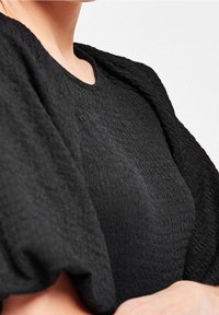 Haut noir texturé avec un col rond et de douces manches bouffantes. Le tissu semble froissé, ajoutant de la profondeur à l'ensemble du design.