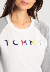 Weißes und graues Raglan-Shirt mit buntem "TOMMY"-Text in verschiedenen Farben, aus weichem Stoff, mit Rundhalsausschnitt und kontrastierenden Ärmeln.