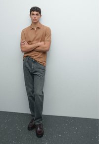 Young man with short hair stands with arms crossed, wearing a brown polo shirt, gray jeans, and dark brown shoes against a light wall.