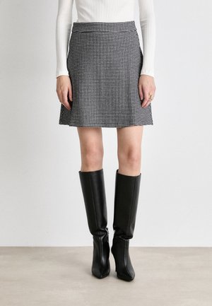 Mujer que lleva una minifalda de pata de gallo en blanco y negro, una camiseta blanca de manga larga con canalé y botas de cuero negras hasta la rodilla, de pie sobre un suelo claro.