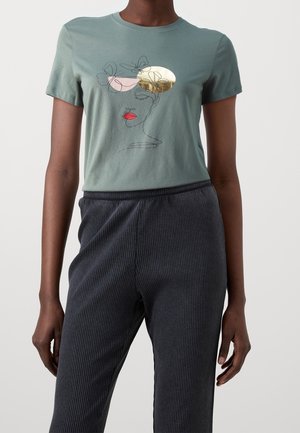 T-shirt vert à manches courtes avec un art linéaire abstrait représentant un visage, des papillons et des formes métalliques dorées et roses, porté avec un pantalon côtelé foncé.