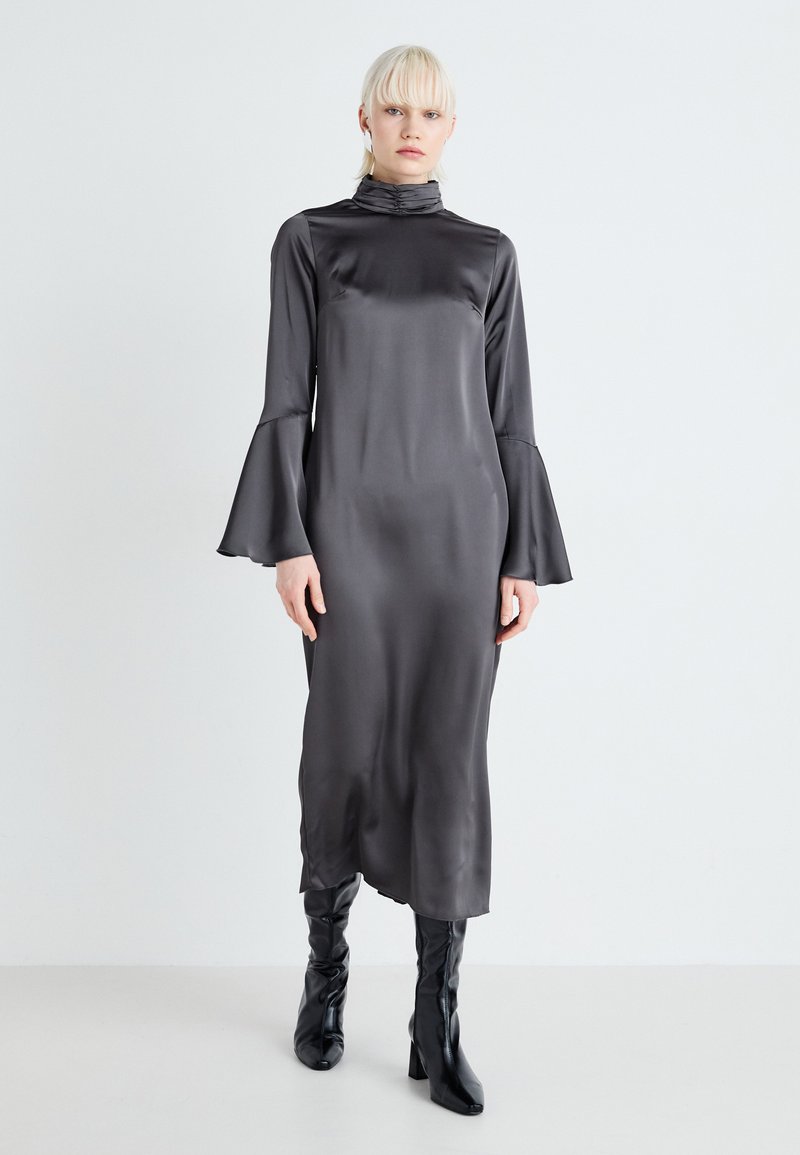 Lang, mørkegrå satin kjole med høy krage og flagrende ermer, bæres med sorte knelange støvler. Glatt tekstur og minimalistisk design.